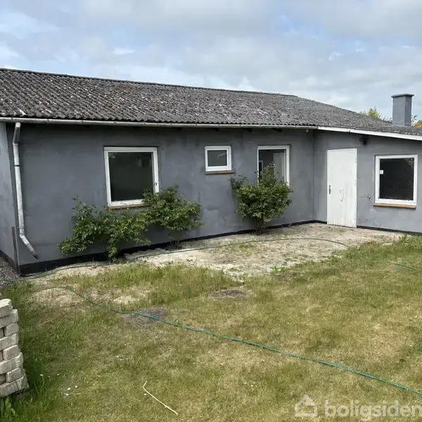 Gråt hus med sorte tagsten står stille i en have. Flere buske vokser tæt på væggen, mens plæner omgiver bygningen. En blå tønde ses ved siden af huset.