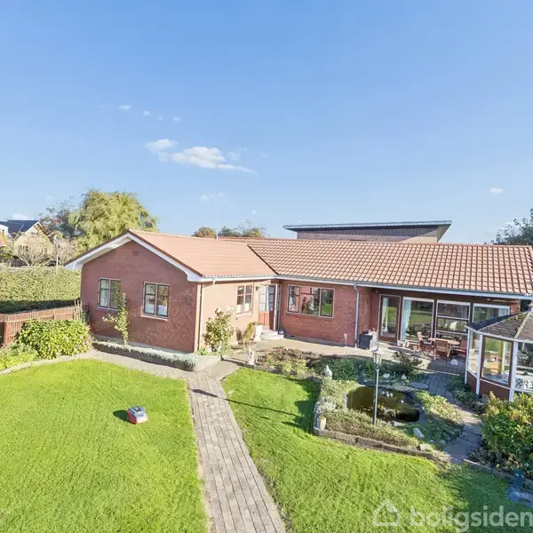 En rød murstensbygning med rødt tegltag ligger midt i en velplejet have. Til højre ses et drivhus. Himmel og andre huse danner baggrund.