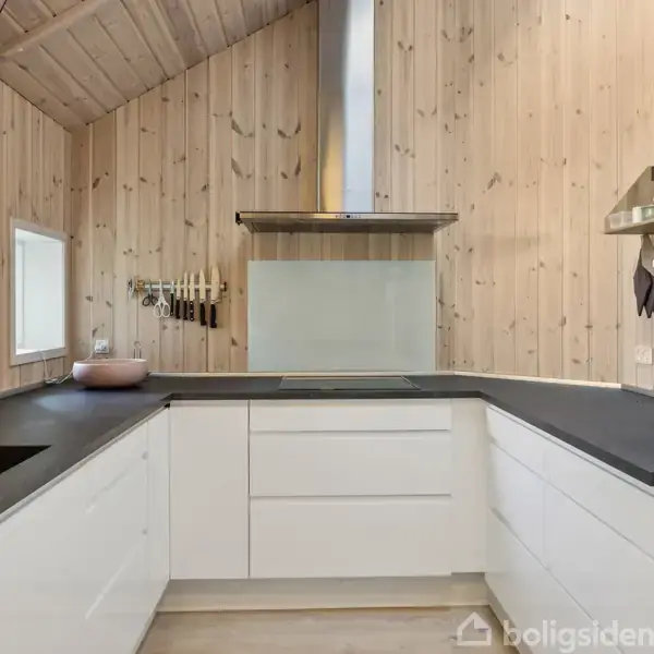 Et moderne køkken med hvide skabe og sorte bordplader. Væggene er beklædt med lyst træ. Indbygget ovn til højre, vindue til venstre, køkkenvask og komfur centralt placeret.