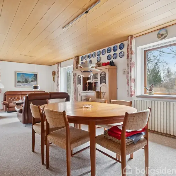 Et spisebord med stole står midt i en stue, omgivet af lænestole og en væghylde med tallerkener. Stuen har store vinduer og et træloft.