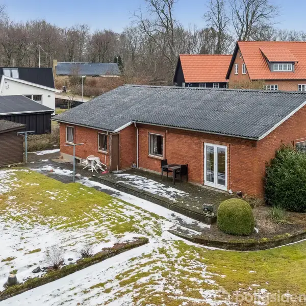 Et rødt murstenshus står stille i en vinterhave med spredt sne, omgivet af andre huse med baggrund af træer og en blå himmel. To borde står på terrassen.