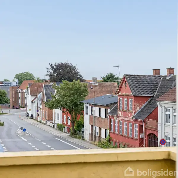 Gaden med farverige huse ses fra en altan. Vinrøde og hvide bygninger står side om side langs vejen, der drejer til venstre, med grønne træer i baggrunden.