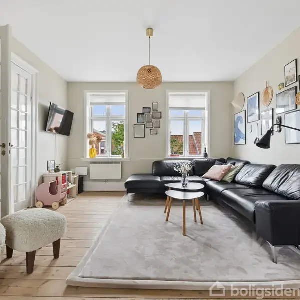 Stue med sort lædersofa langs væg, sofabord i træ på tæppe, og væg med kunstværker. Et fjernsyn hænger over for sofaen. Værelset oplyses af to store vinduer.