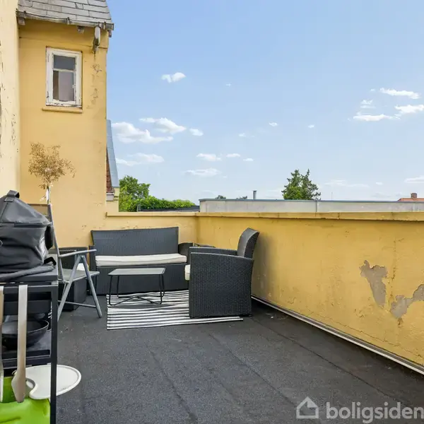 Terrasse med to sofaer og et bord, grill på et lille bord med redskaber. Omgivet af gule vægge med synlig afskalning og udsigt over hustage og træer.