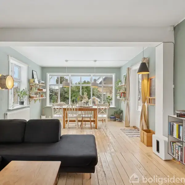 Sort sofa står i et lyst rum med trægulve, hvor et spisebord er placeret i en oplyst karnap. Bøger og dekorative elementer pryder reolerne langs væggene.