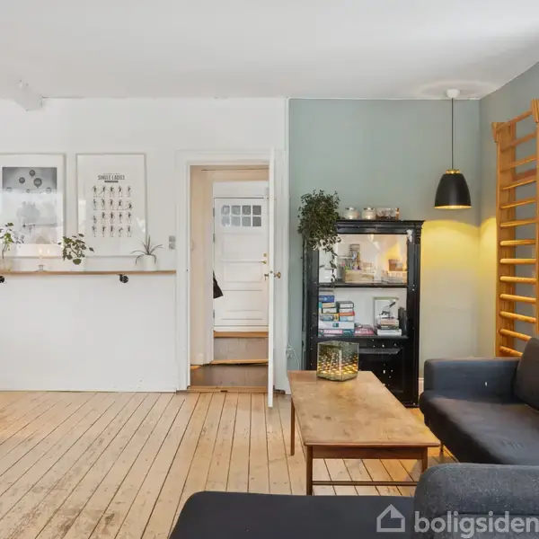 En stue med trægulv og grå vægge. En sort sofa og et træbord står ved vinduet. En reol og billeder hænger på væggen. En lampe lyser rummet.