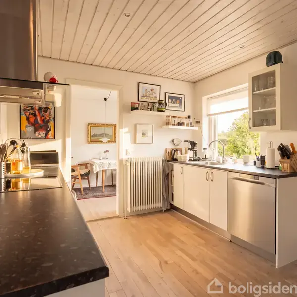 Køkken med sorte bordplader og hvide skabe langs væggen. Rum lyses op af dagslys fra stort vindue. Omgivelser inkluderer spisestue i baggrunden med malerier på væggene.