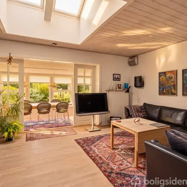 Stue med sort lædersofa og fladskærms-TV på mørkt træbord. Stueplante og pejsebrænde til højre. Store vinduer og dør fører til terrasse med træstole, omgivet af grønne planter. Skjult loft med ovenlysvinduer.