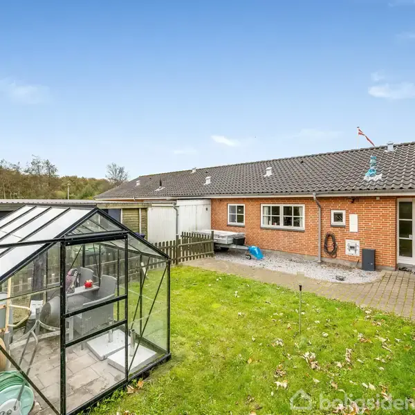 Drivhus står på en græsplæne i en baghave. Ved siden af ses en rød murstensbygning med vinduer og en dør, omgivet af en flisebelagt sti og haveudstyr.