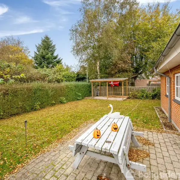 En udendørs bænk med to udskårne græskar står på en flisebelagt terrasse ved et rødt murstenshus omgivet af grønt græs, hække og træer. En stang ses i baggrunden.