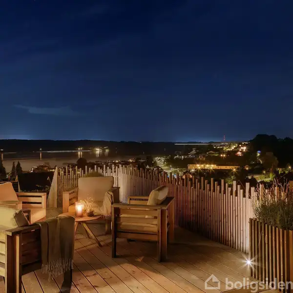 Terrasse med træmøbler og planter. Et bord har to vinglas og en vinkøler. Udsigt til en sø og skove under en blå himmel med skyer.