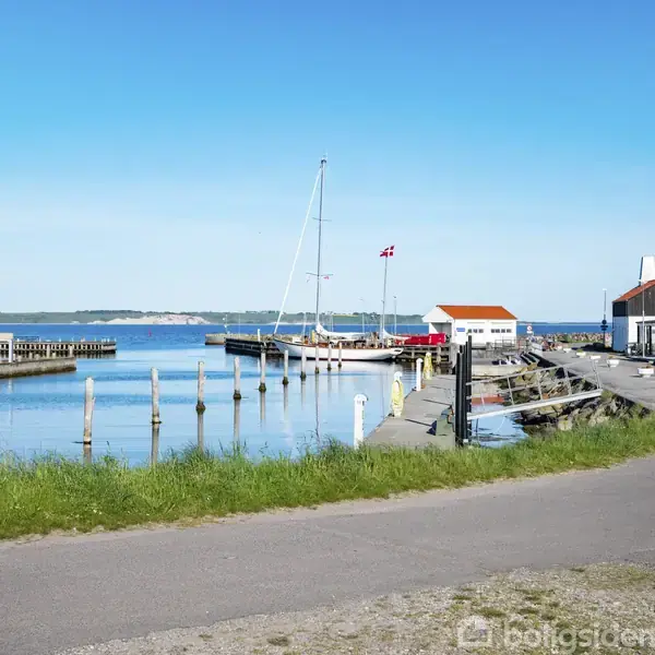 En lystbåd ligger i en havn med en dansk flag på masten. Omgivet af små bygninger ved en rolig kystlinje under en klar blå himmel.