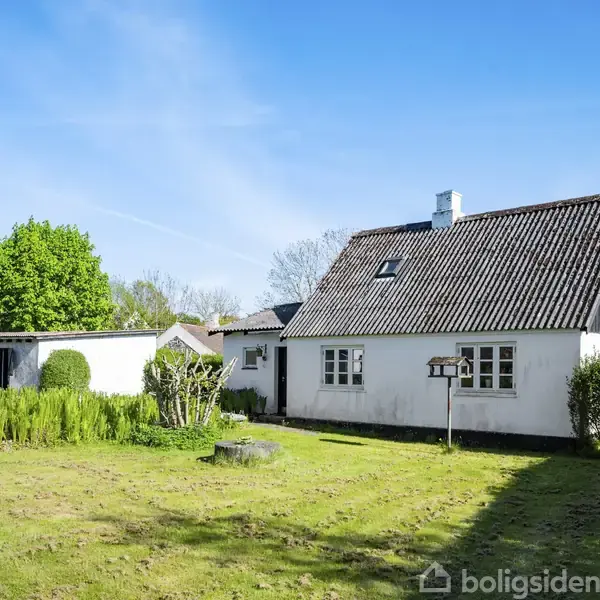 Et hvidt hus med skråt tag står i en grøn have. Træer og buske omgiver huset under en klar blå himmel.