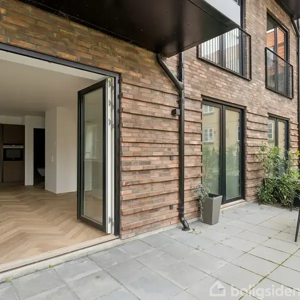 Åben dobbeltdør fører fra en moderne lejlighed med trægulv til en flisebelagt terrasse. Tre stabelbare stole og et rundt bord er placeret ved siden af en række grønne planter.
