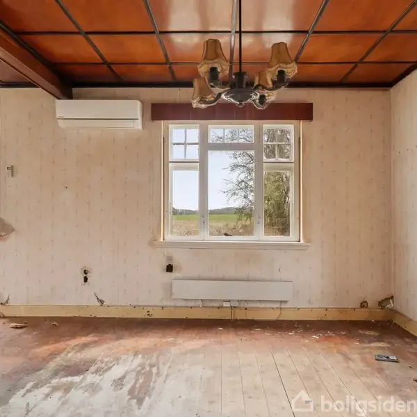 Et tomt værelse med trælaminatgulv og brune lofter. En hængende loftslampe er centralt placeret. Vinduerne viser en udsigt over grønt landskab. Radiator og aircondition er på væggen.