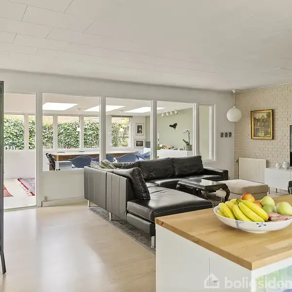 Sofa i sort læder står i en stue med en fladskærm på væggen. I forgrunden ses en skål med frugt på en køkkenø. Store vinduer giver udsigt til haven.