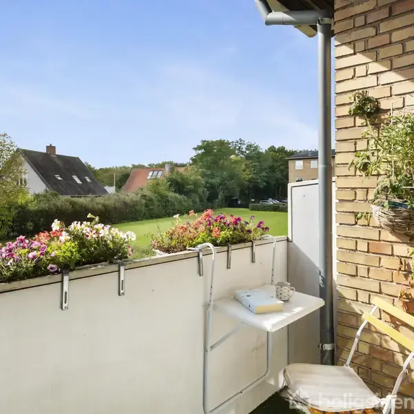 En lille altan med blomsterkasser fulde af farverige blomster. Et bord og en stol er arrangeret til afslapning. I baggrunden ses en græsplæne og huse.