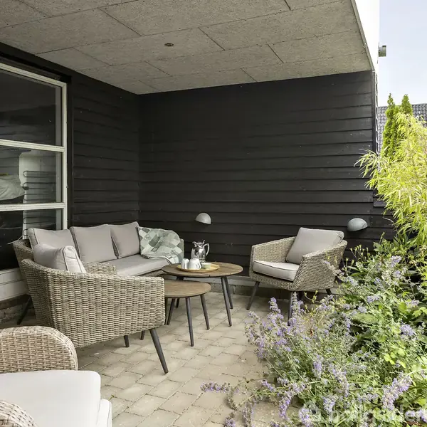 Terrassemøbler af flet står ordnet i en hyggelig krog. De omkranser et lille bord med planter i forgrunden. Mørke træpaneler udgør væggene i den beskyttede udendørsområde.