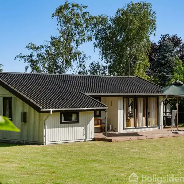 Et enkelt hvidt sommerhus med sort tag står på en velplejet græsplæne omgivet af træer og buske under en blå himmel.