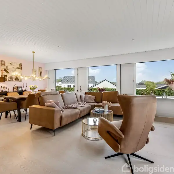 Stue med to sofaer og lænestol centreret omkring et rundt sofabord; et spisebord og stole i baggrunden. Store vinduer giver udsigt til haver og huse udenfor.