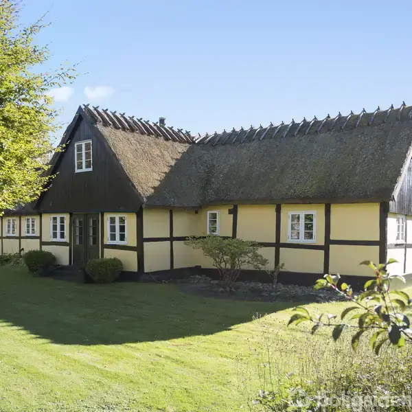 Bindingsværkshus med stråtag står stille i en frodig have med grønne træer og buske, omgivet af en velplejet græsplæne under en klar blå himmel.