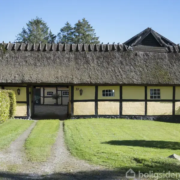 Et bindingsværkshus med stråtag står i en velholdt have. Indkørselen består af grus, og en tæt hæk omkranser området. Nummeret "29" ses ved indgangen.