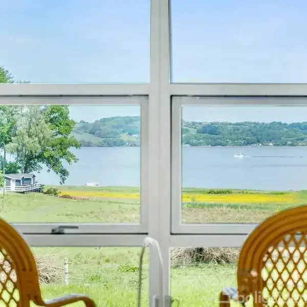 To stole står vendt mod et stort vindue med udsigt til en sø eller fjord og grønne marker. I baggrunden ses træer og en båd på vandet.