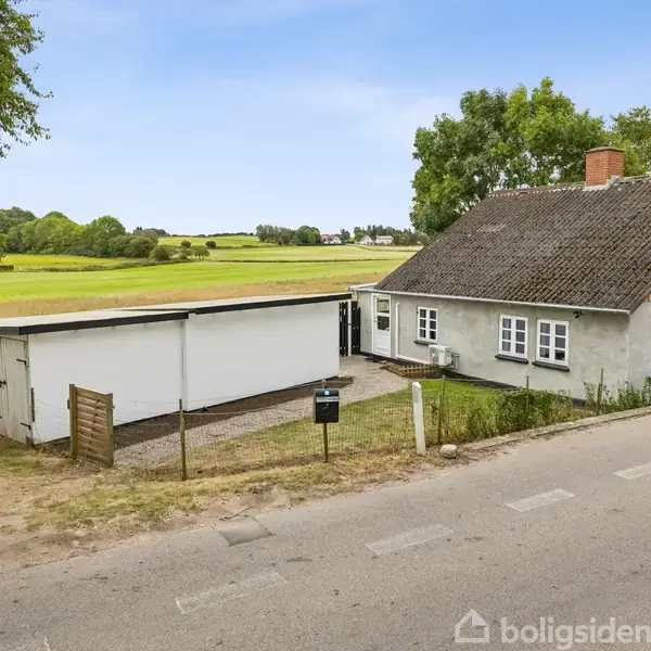 En lille gråmalet hus bygget med mursten og et tilhørende hvidt skur står ved en landevej. Baggrunden består af grønne marker og træer under en klar blå himmel.