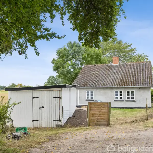 En hvidmalet landsbyhus med brun tag, placeret ved en vej. Foran huset står en træskur. Omgivet af grønne træer og åbne marker under en blå himmel.