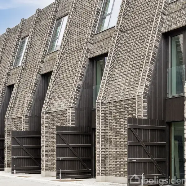 Bygning med skrå, strukturerede murstensfacader; store vinduer integreret i designet. Set i række, skaber en moderne boligstil i et bymiljø.