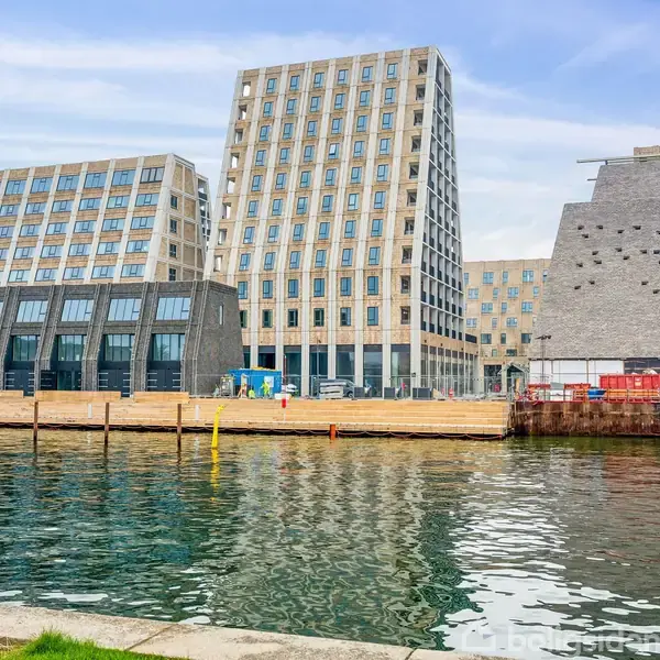 Moderne bygning med store vinduer og skrånende facader står ved en havnefront. Vandet i forgrunden reflekterer strukturen, omgivet af træmoler og himmel med lette skyer.