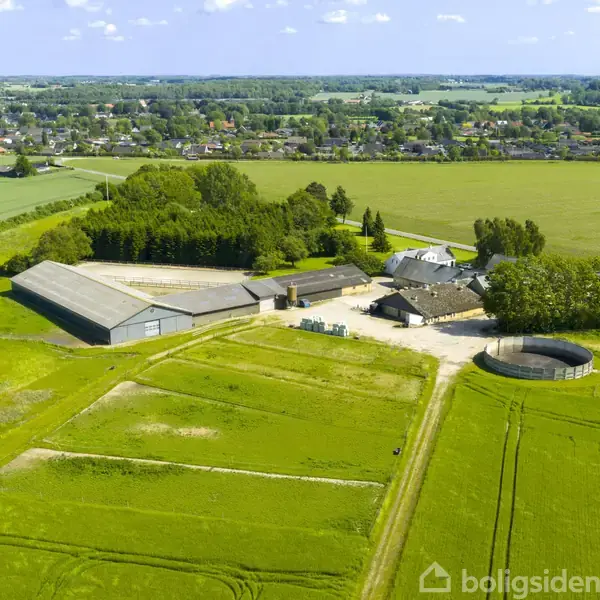 Gårdkompleks omgivet af grønne marker, hvide bygninger og træer samt strukturer til opbevaring. I baggrunden ses en landsby og flere marker under en blå himmel med få skyer.