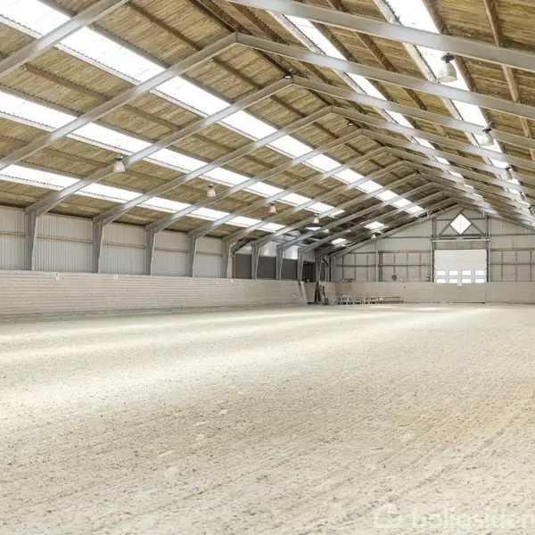 Rideskolens indendørs arena med højt træloft og sanddækket gulv, oplyst af flere rækker ovenlysvinduer. En række stole står ved den fjerne væg. Ingen tekst fundet.