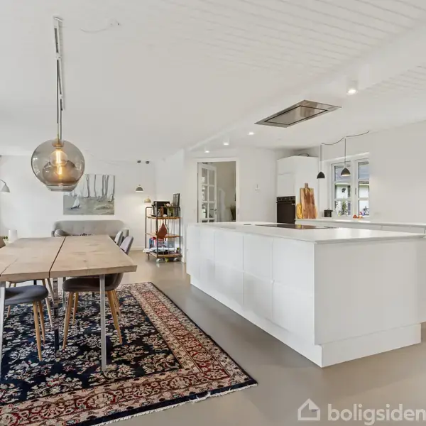 Et moderne køkken og spisestue har et langt træbord på et mønstret tæppe. Køkkenet har en hvid ø og hængende lamper. Rummet er lyst med minimalistisk indretning.
