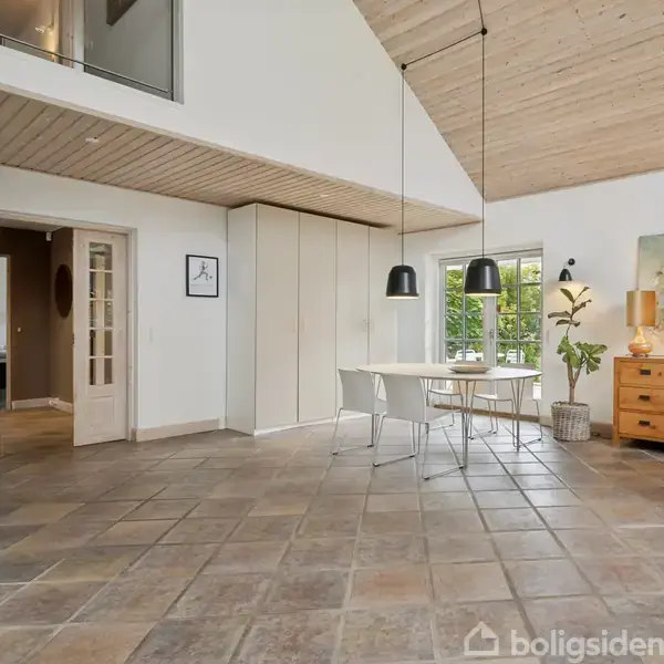 Et hvidt spisebord med to stole står i et lyst rum med træloft og flisegulv. Der er to sorte pendellamper, et vindue, en plante, og en kommode med lampe.