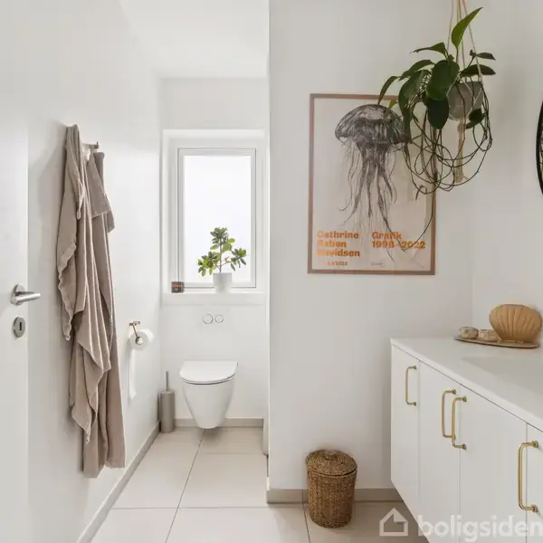Toilet i lyst badeværelse med hængende planter og vægkunst. Omgivet af hvid indretning, håndvask med spejl, håndklæde på væggen. Vindue giver naturligt lys.