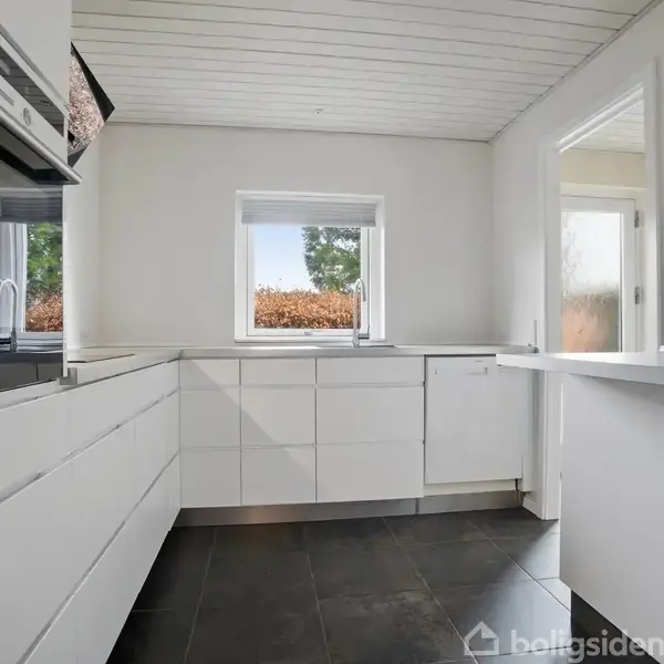 Hvidt køkken med moderne skabslåger, ovn og vask. Rummet er lyst med et stort vindue, der har udsigt til en hæk. Mørkt flisegulv og hvide vægge.