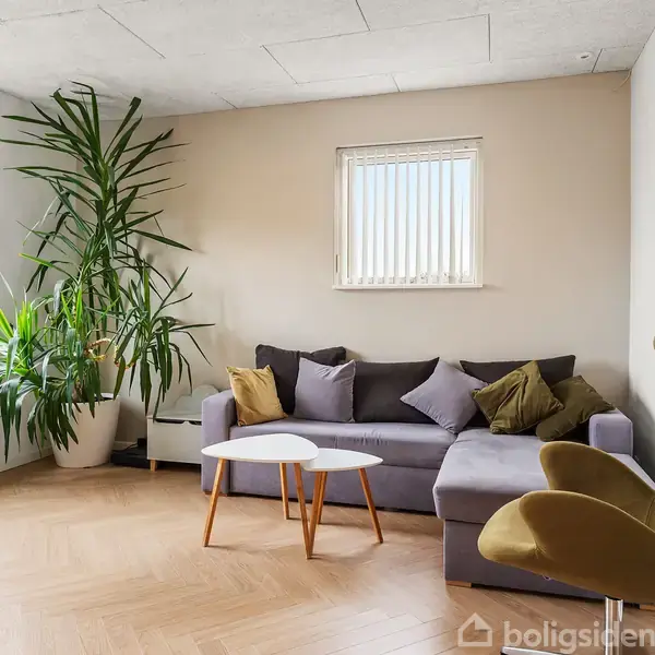 Stue med en grå sofa fyldt med puder og en grøn lænestol. To lave borde står foran sofaen. Store grønne planter i hjørnet ved siden af et vindue med persienner.