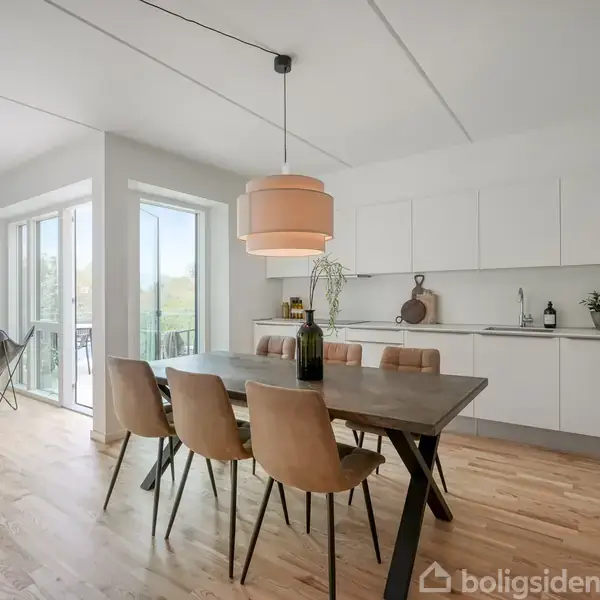 Spisebord med seks stole står i en moderne, lyst køkken-alrum. En stor pendellampe hænger over bordet. Der er adgang til en balkon gennem store glasdøre, og stuen ses i baggrunden.