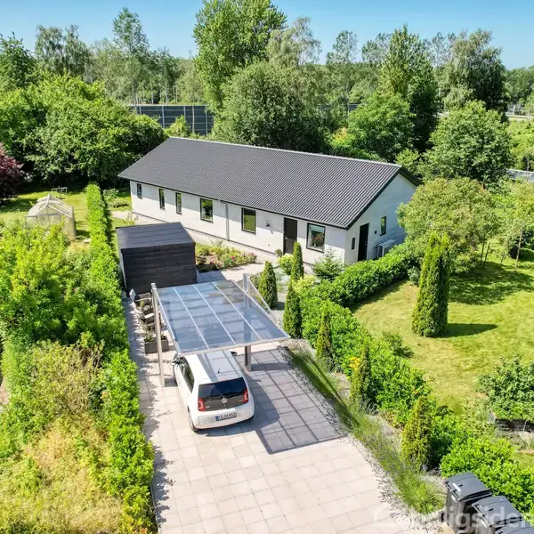Hus med sort tag, omkranset af frodig have. Bil under carport på flisebelagt indkørsel. Træer og buske omkranser området, skaber en privat atmosfære.