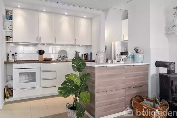 Køkken med hvide skabe og indbygget ovn, pejs ved siden af trækassette, grøn plante i potte; køkkenredskaber på bordplade. Lyst interiør med moderne detaljer.