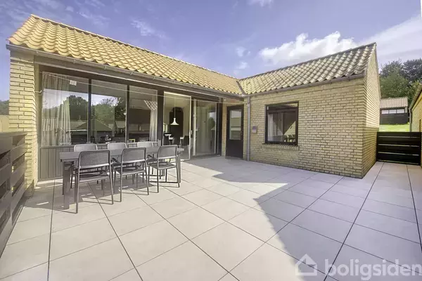 Et gult murstenshus med glasfacader og skrå tag. Der står et udendørs spisebord med stole på en flisebelagt terrasse under blå himmel omgivet af grønne træer.