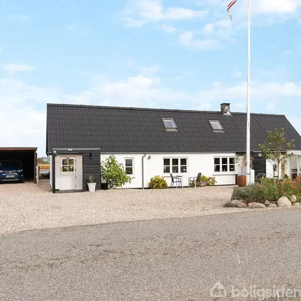 Et hvidt hus med sort tag står stille på en grusplads ved siden af en carport med to biler. En flagstang ses i forgrunden mod en blå himmel.