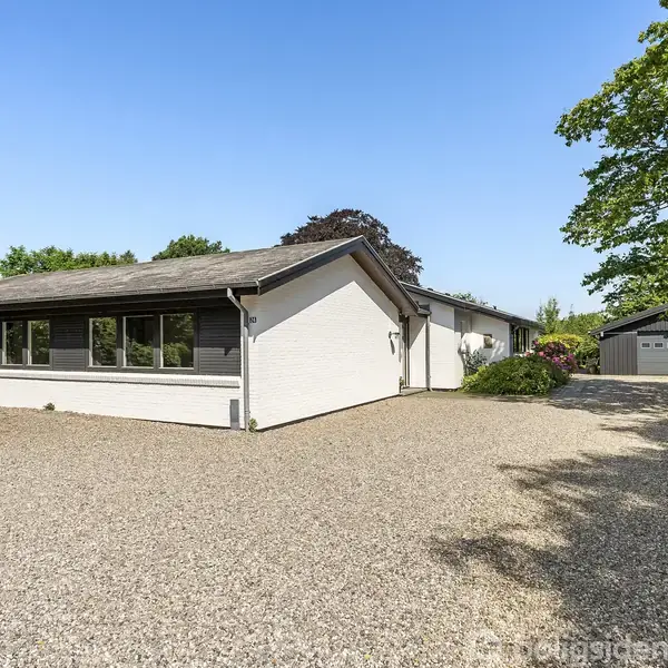 Et hvidt, en-etagers hus står stille omgivet af en grusbelagt forplads og frodige træer, med en garage i baggrunden. Klar blå himmel over stedet.