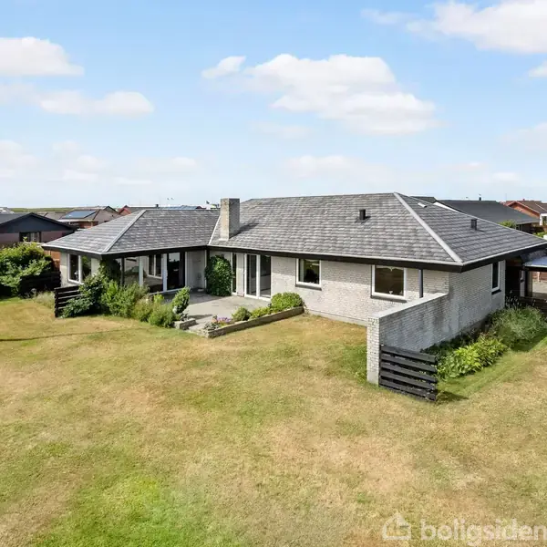 En rummelig, ensomtliggende bungalow står midt i en stor, velholdt græsplæne med hegn og høj flagstang. Omkringliggende parcelhushuse skaber en rolig forstadsatmosfære under en lys, delvis skytteklar himmel.
