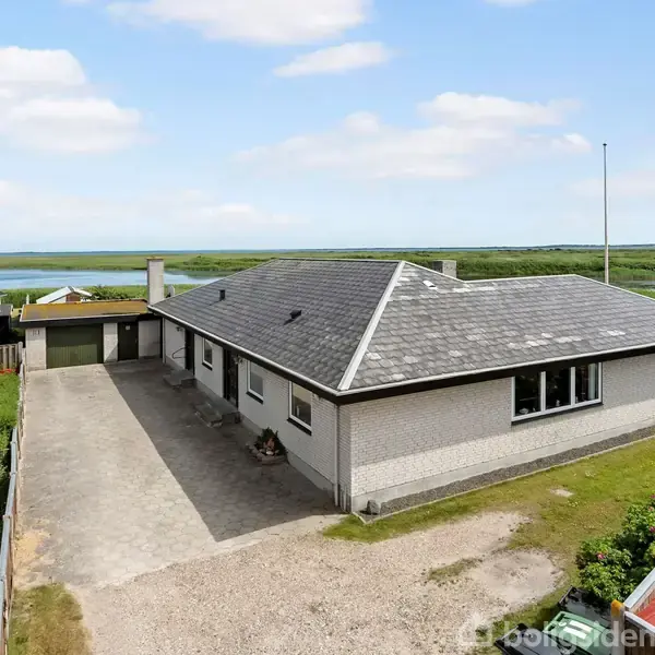 Et hvidt hus med gråt tag står stille. Det er omgivet af en flisebelagt indkørsel og grønne haver. I baggrunden er der en sø og blå himmel.