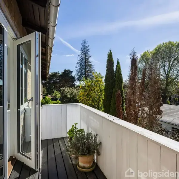 Åben altandør til en træbeklædt altan med planter i krukker. Omgivet af træer under en klar blå himmel i et roligt boligområde.