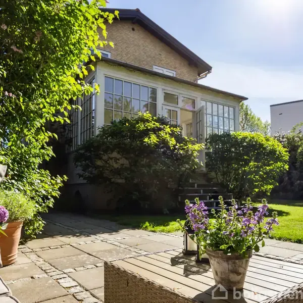 En stenbelagt terrasse med blomsterkrukke og havemøbler foran en murstensbygning med store vinduer. Omgivet af grønne buske og græsplæne i solrige udendørs omgivelser.