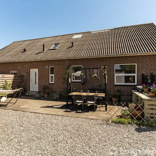 Rødt murstenshus med stor terrasse med spisebord og stole. Omkringliggende have med blomster og planter i krukker og bede under en blå himmel. Ingen tekst til stede.