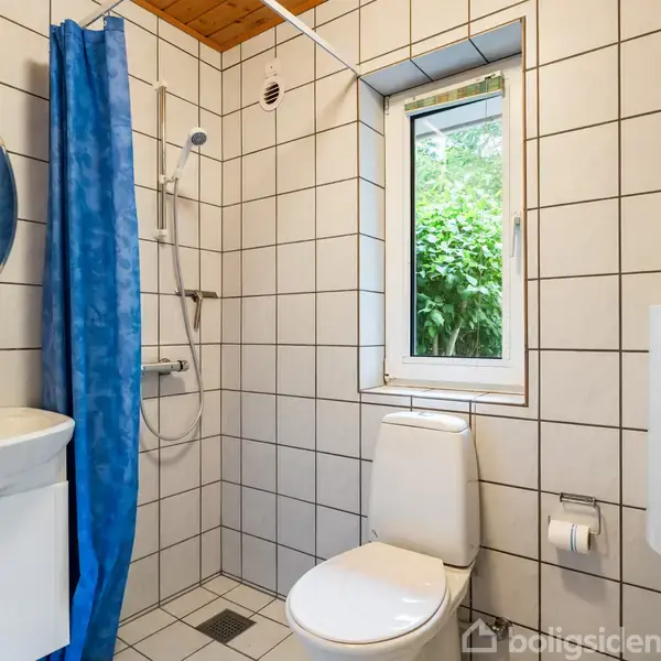 Et lille badeværelse med hvidkaklede vægge har et toilet, en håndvask med rundt spejl, og en bruser med blå forhæng. Et vindue med udsigt til grønne buske giver naturligt lys.
